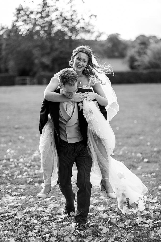 3 of 3 – black and white photo of laughing groom giving his smiling bride a piggy-back walking through park