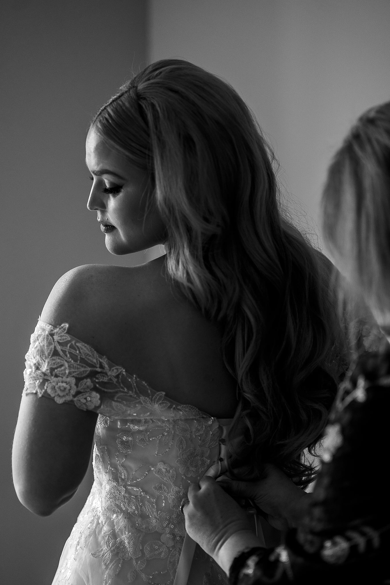 Black and white photo of elegant bride with long hair having her wedding dress fastened at the back, while looking over her shoulder