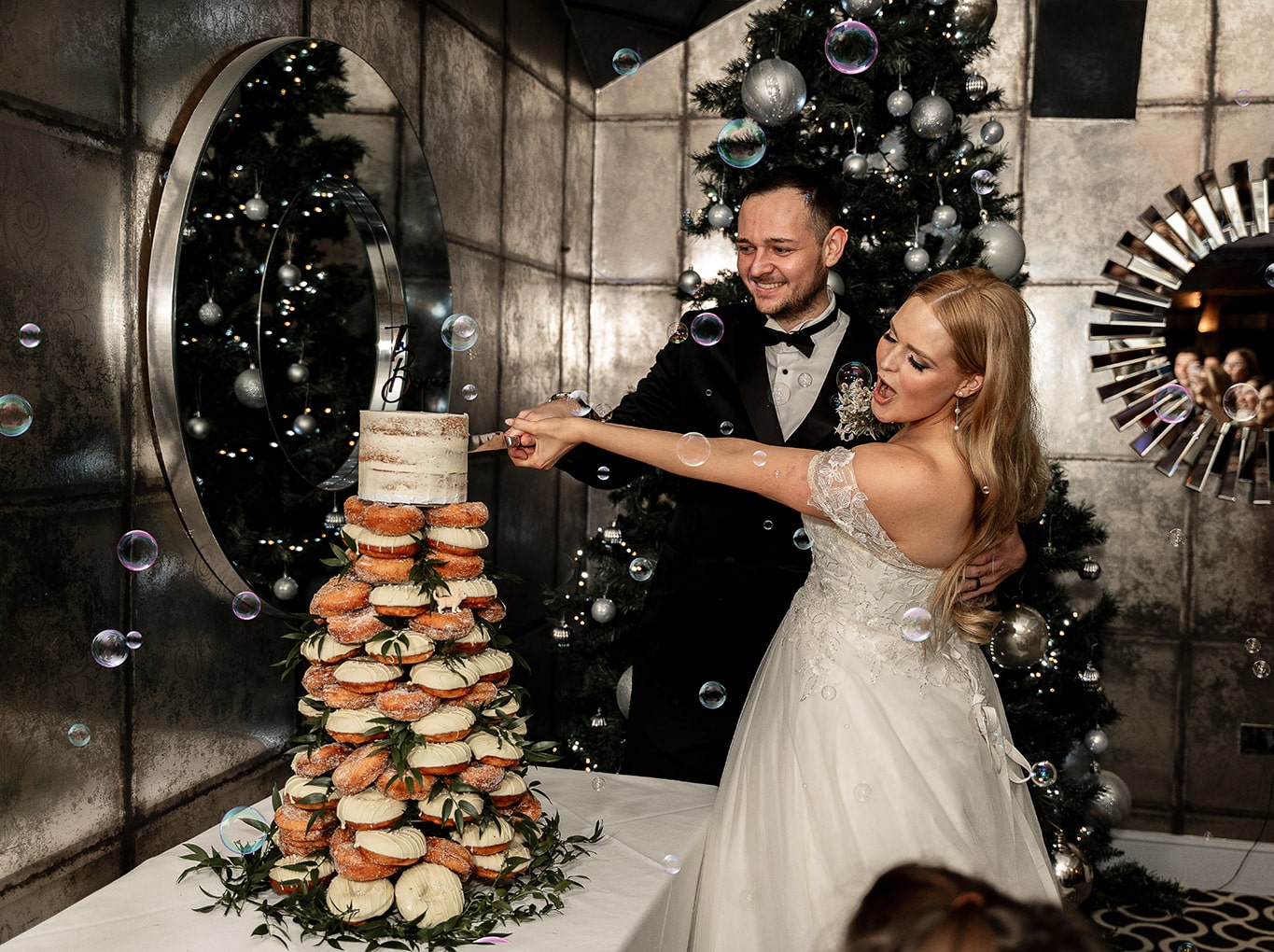 Happy fun photo of bride and groom cutting their doughnut tower wedding cake while bubbles are released. Various Christmas decorations and mirrors in background