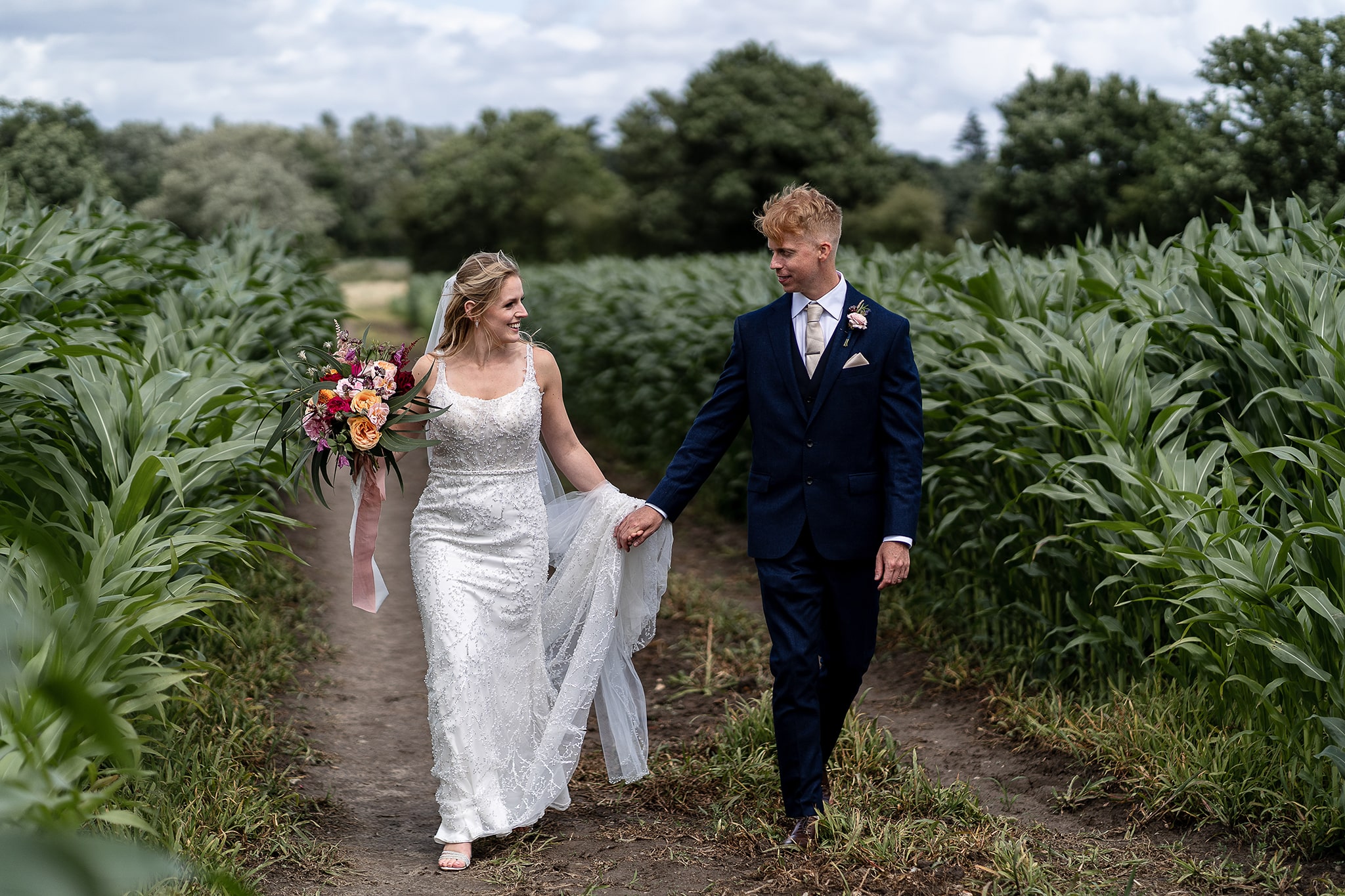 Bride in white wedding dress and groom in contemporary blue suite walking down path through cornfield in Hampshire