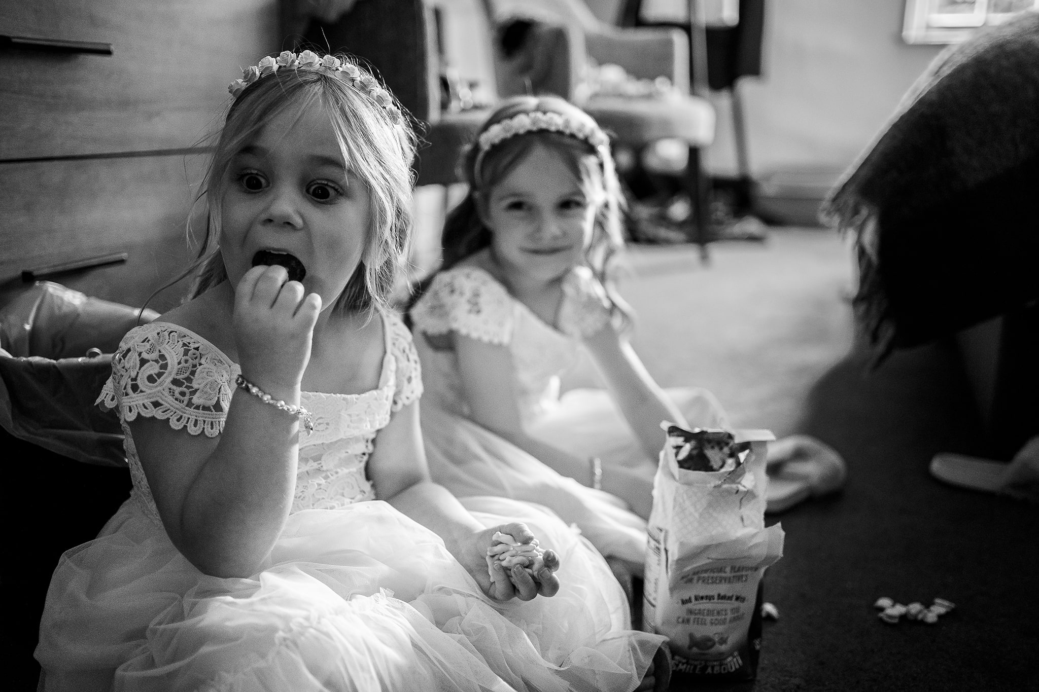 Great documentary style black and white photo taken by professional wedding photographer Emma Seaney of a pair of cute little girl bridesmaids sitting on the floor cheekily stuffing their faces with mini fish-shaped cheesy crackers
